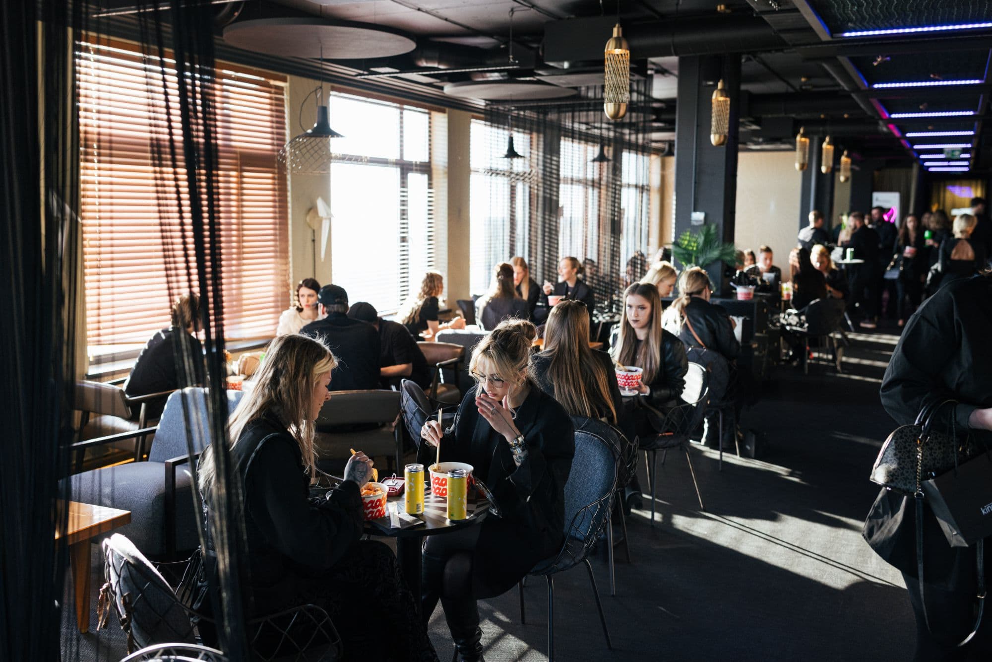 A sunlit event lounge full of people talking and eating at small tables.