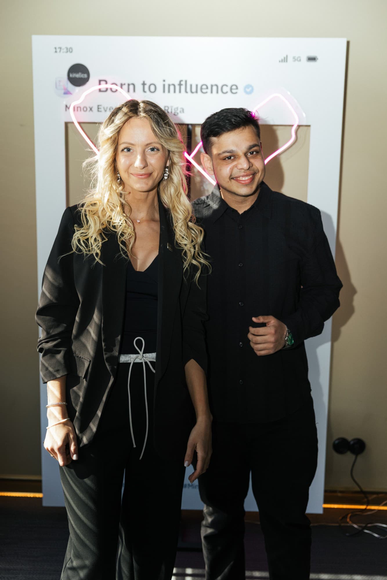 Two people smiling in front of a white "Born to influence" photo frame with a pink neon heart.